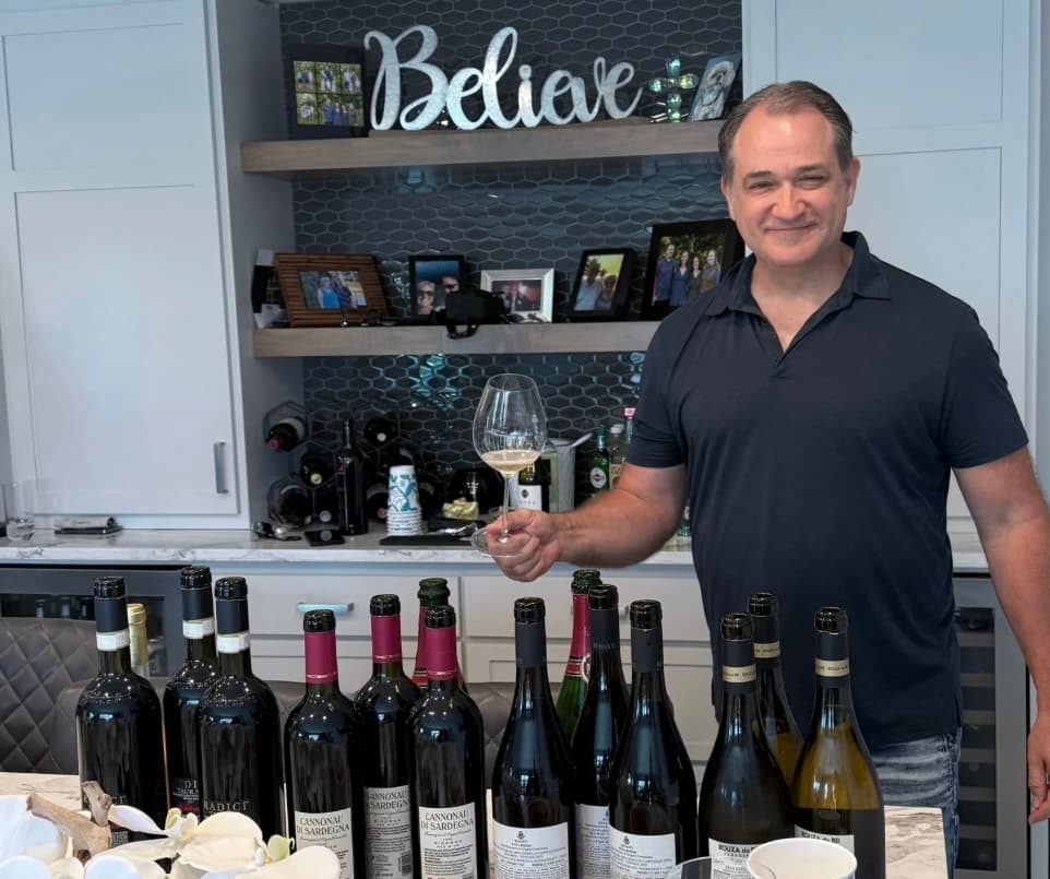 Smiling man holds a glass of wine behind a counter filled with wine bottles.
