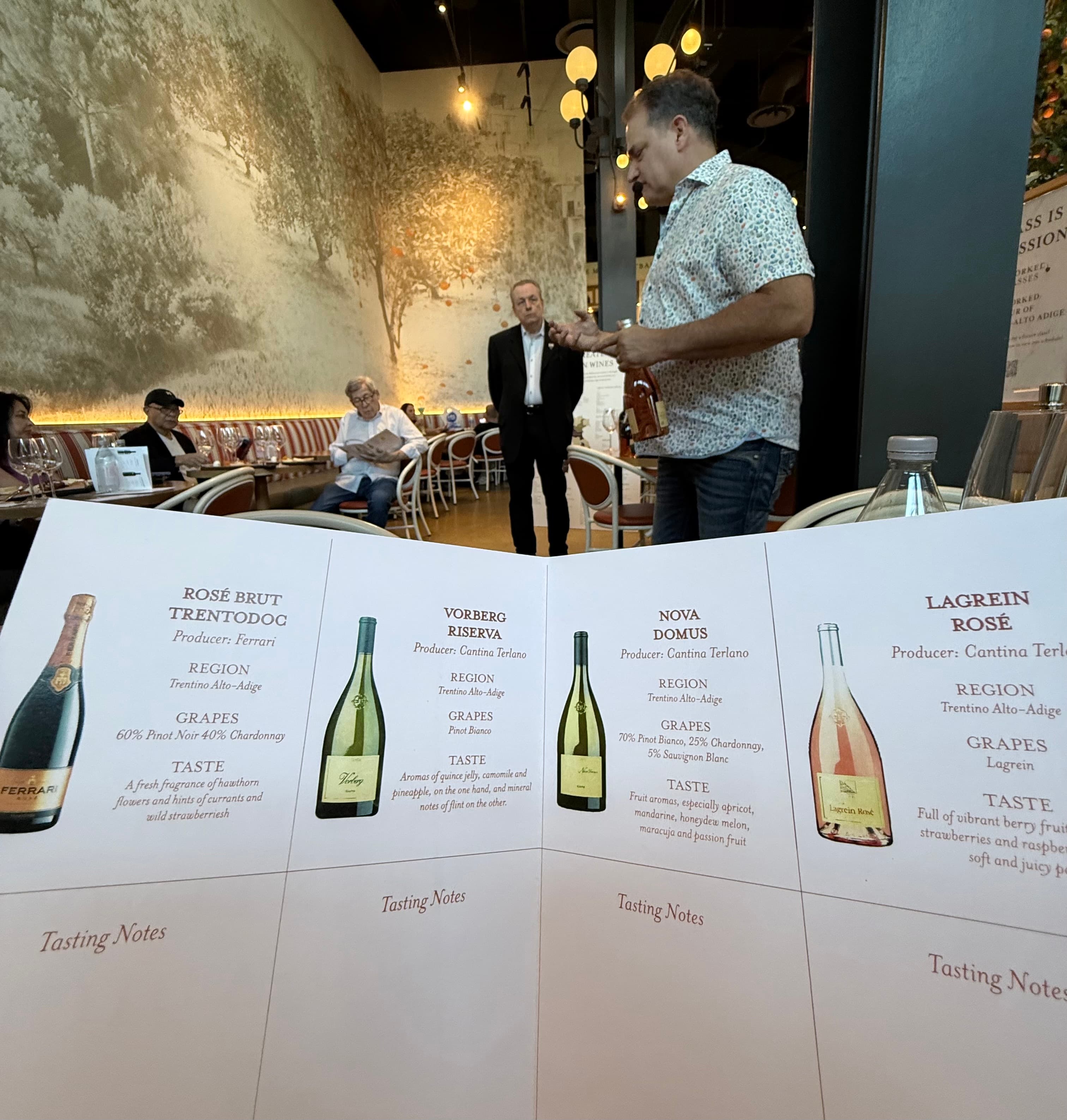 Wine tasting menu in foreground with a man presenting a bottle to restaurant guests.