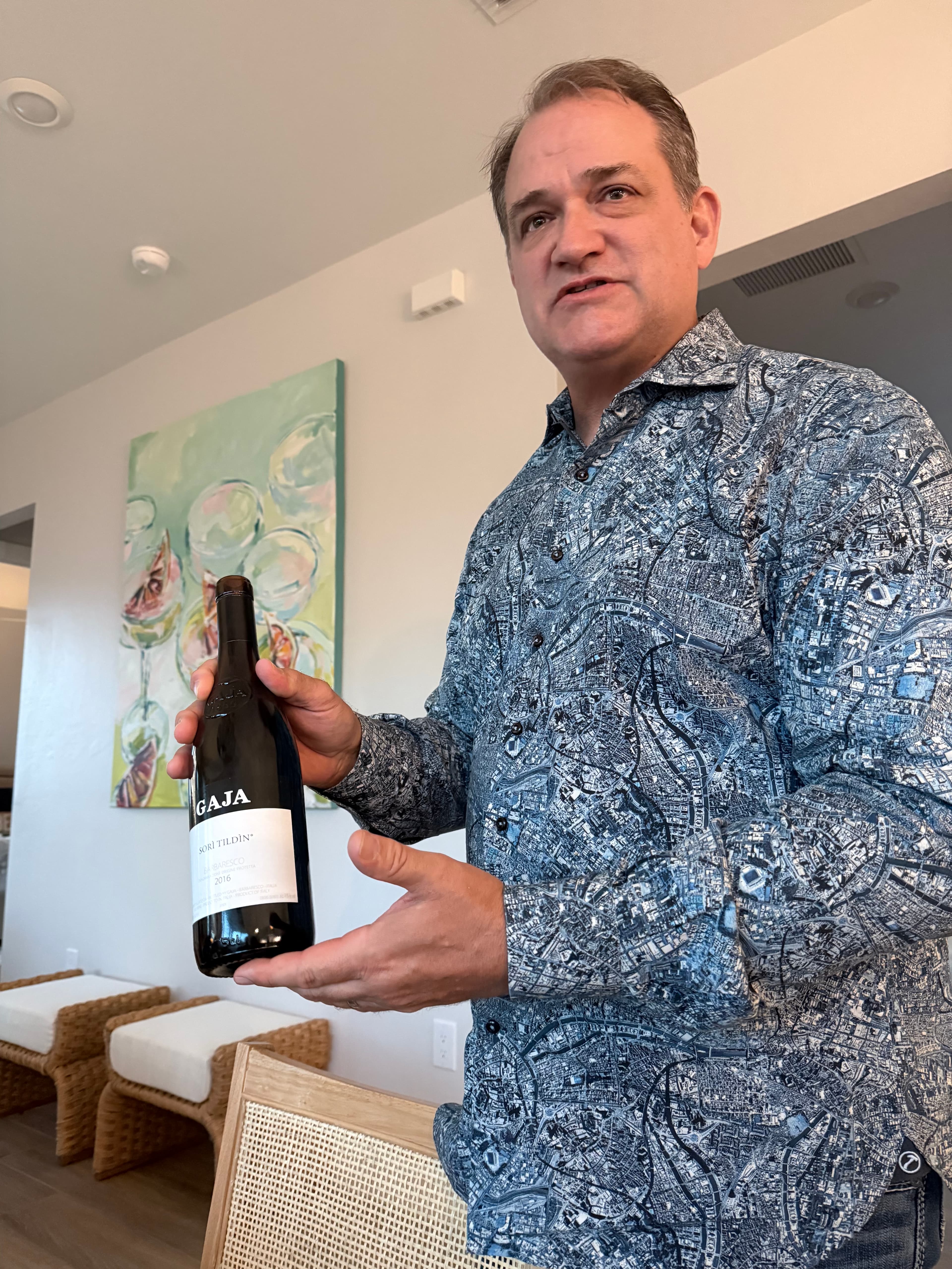 Man in a blue map-patterned shirt holding a bottle of Gaja Sori Tildin wine.