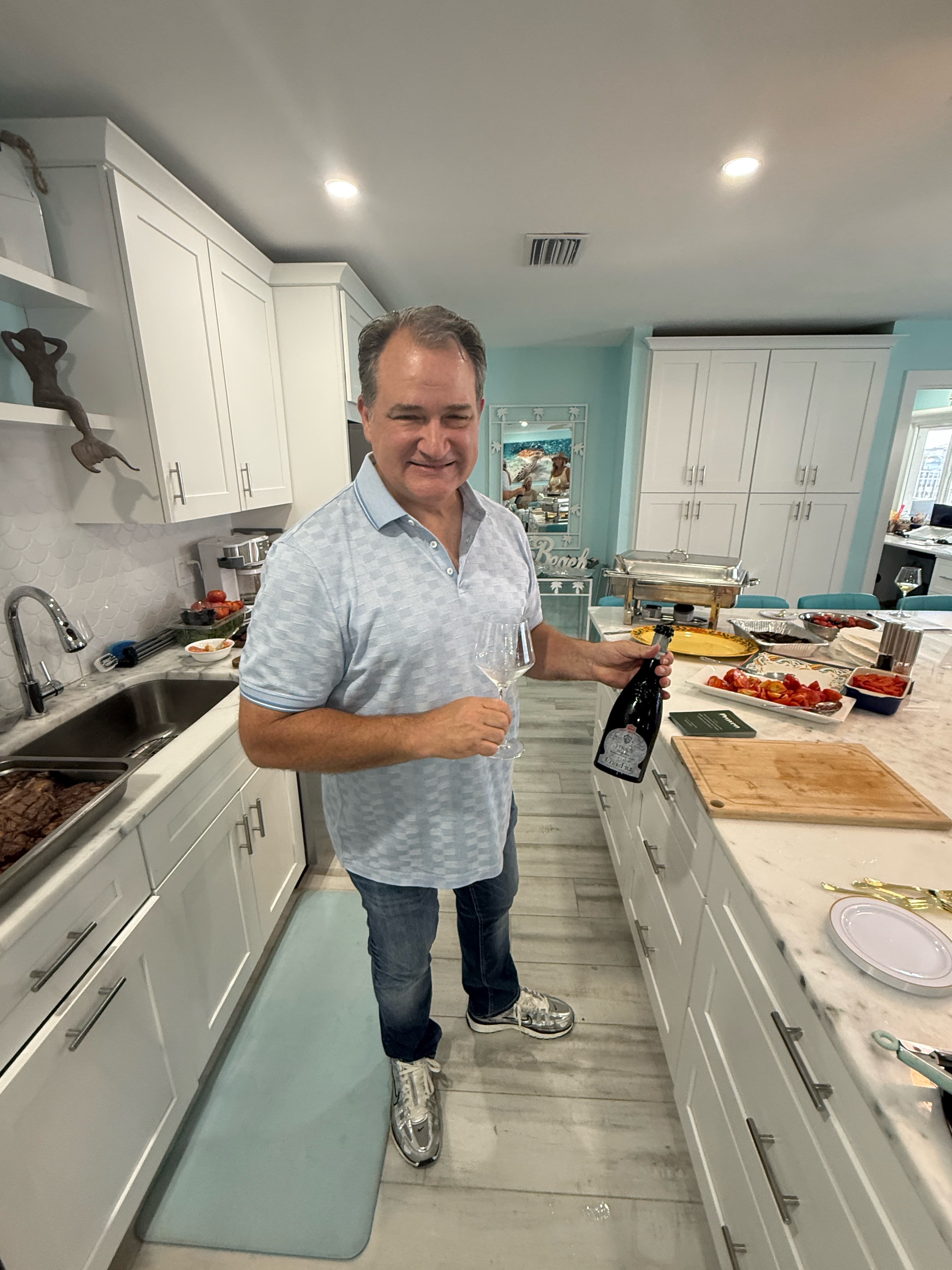 Man in a blue shirt holding a wine bottle and glass in a white kitchen.
